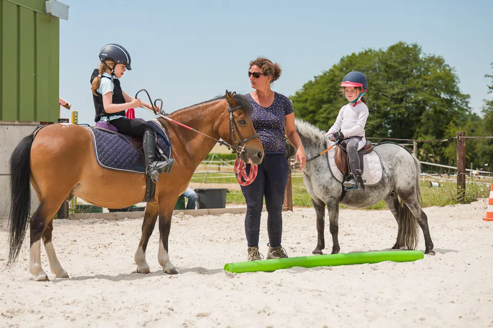 Leçon dans la carrière : la monitrice s’adresse à deux jeunes cavalières, sur un poney bai et un poney gris, devant une barre posée au sol.